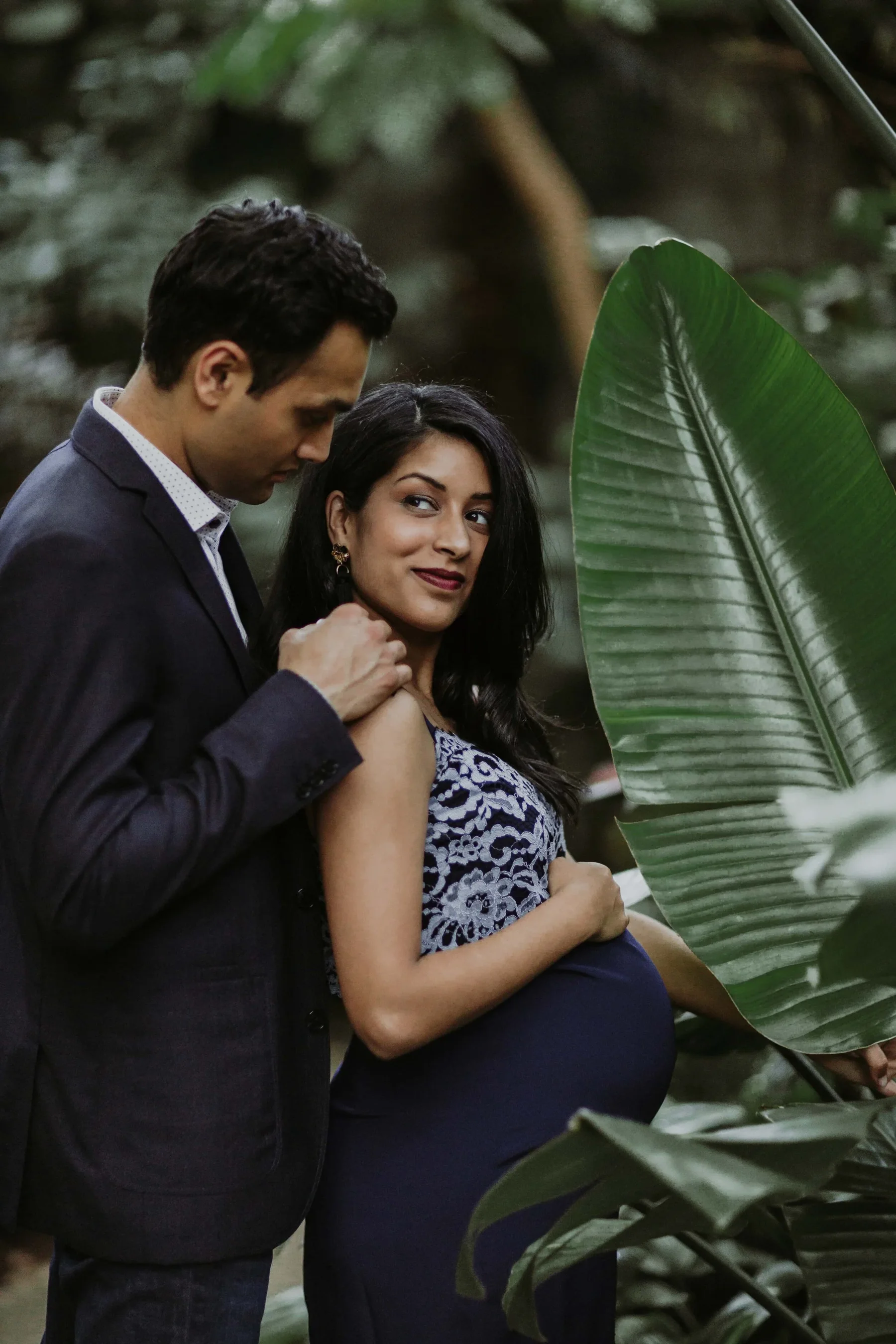 A man in a dark suit gently touches a pregnant woman's shoulder as she stands among large green leaves, looking at the camera with a soft smile in a lush, forest-like setting.