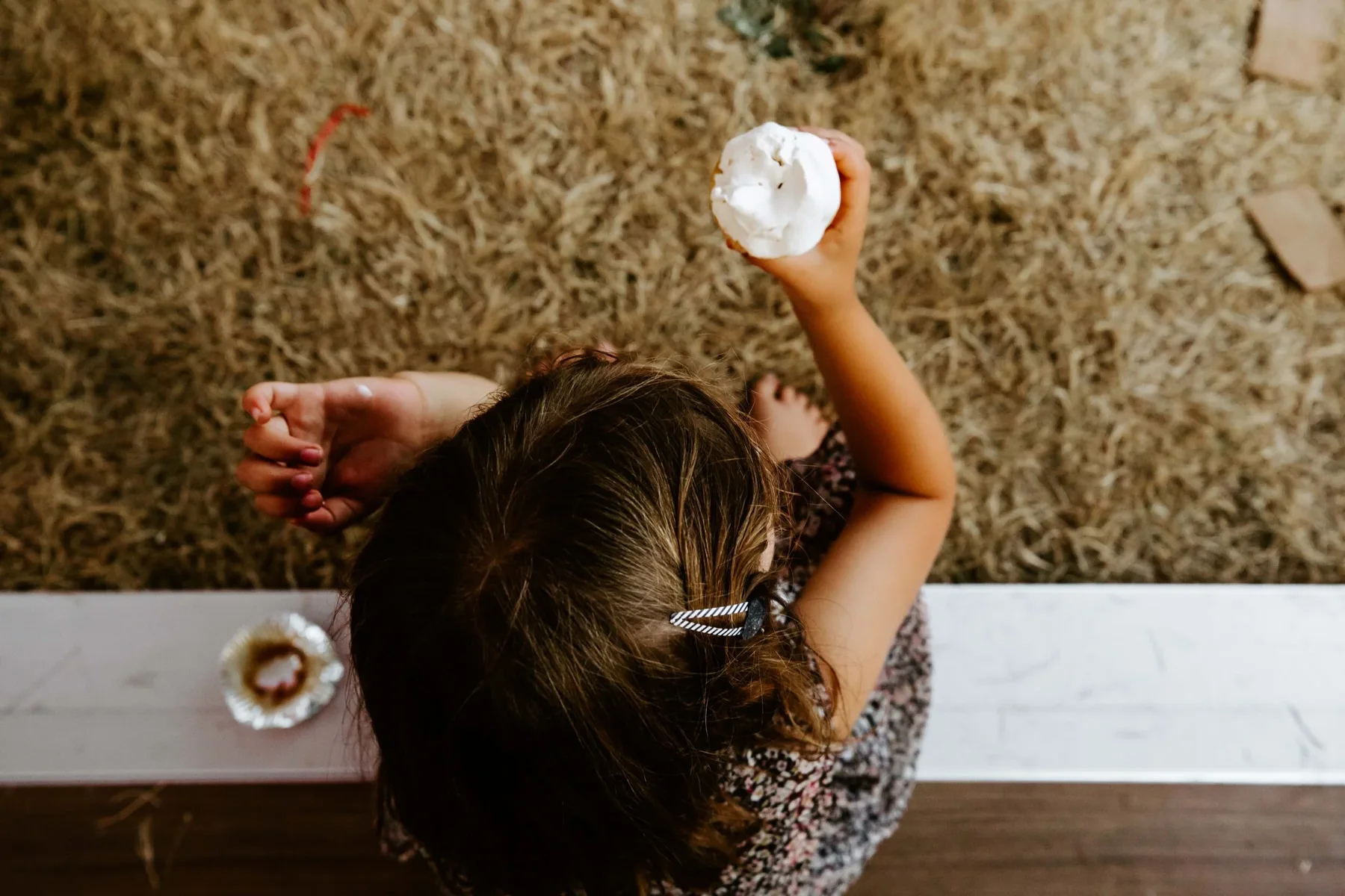 A young girl sitting on the ground holding a half-eaten cupcake with white frosting, viewed from above.