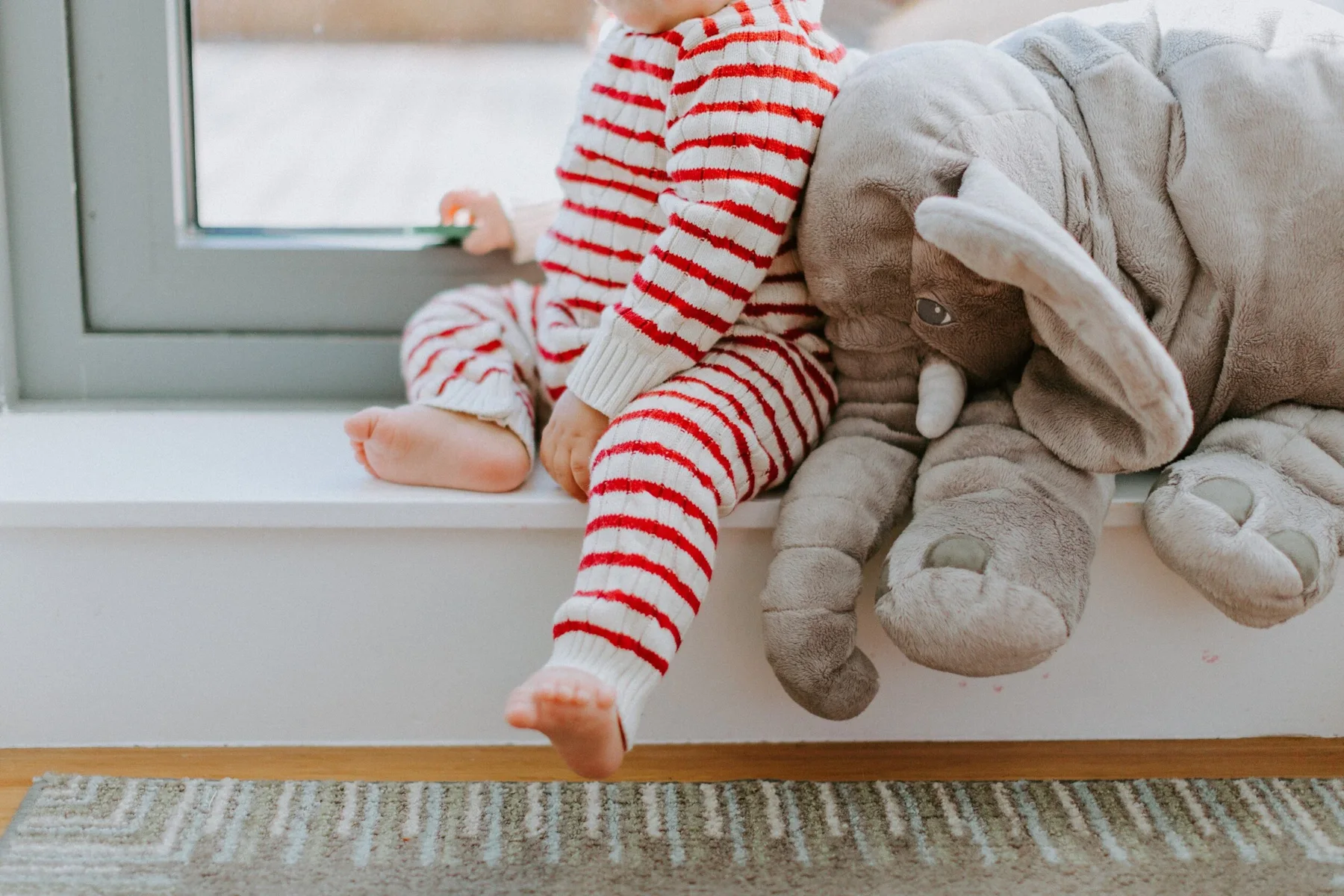 Child sitting on a window sill wearing red and white striped pajamas, with a large gray elephant stuffed animal beside them.