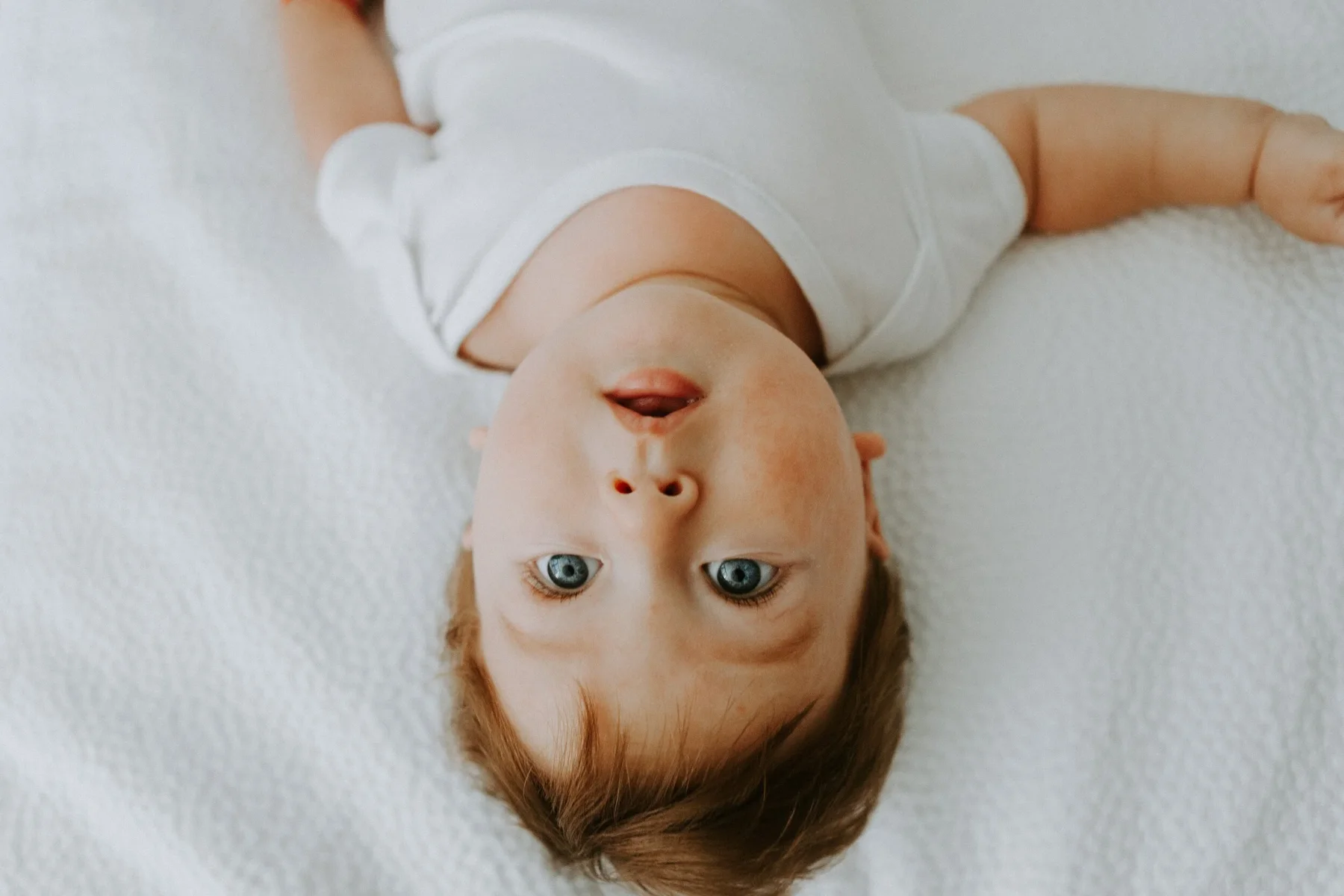 A baby with blue eyes lying on a white textured blanket, wearing a white sleeveless onesie, looking directly at the camera.