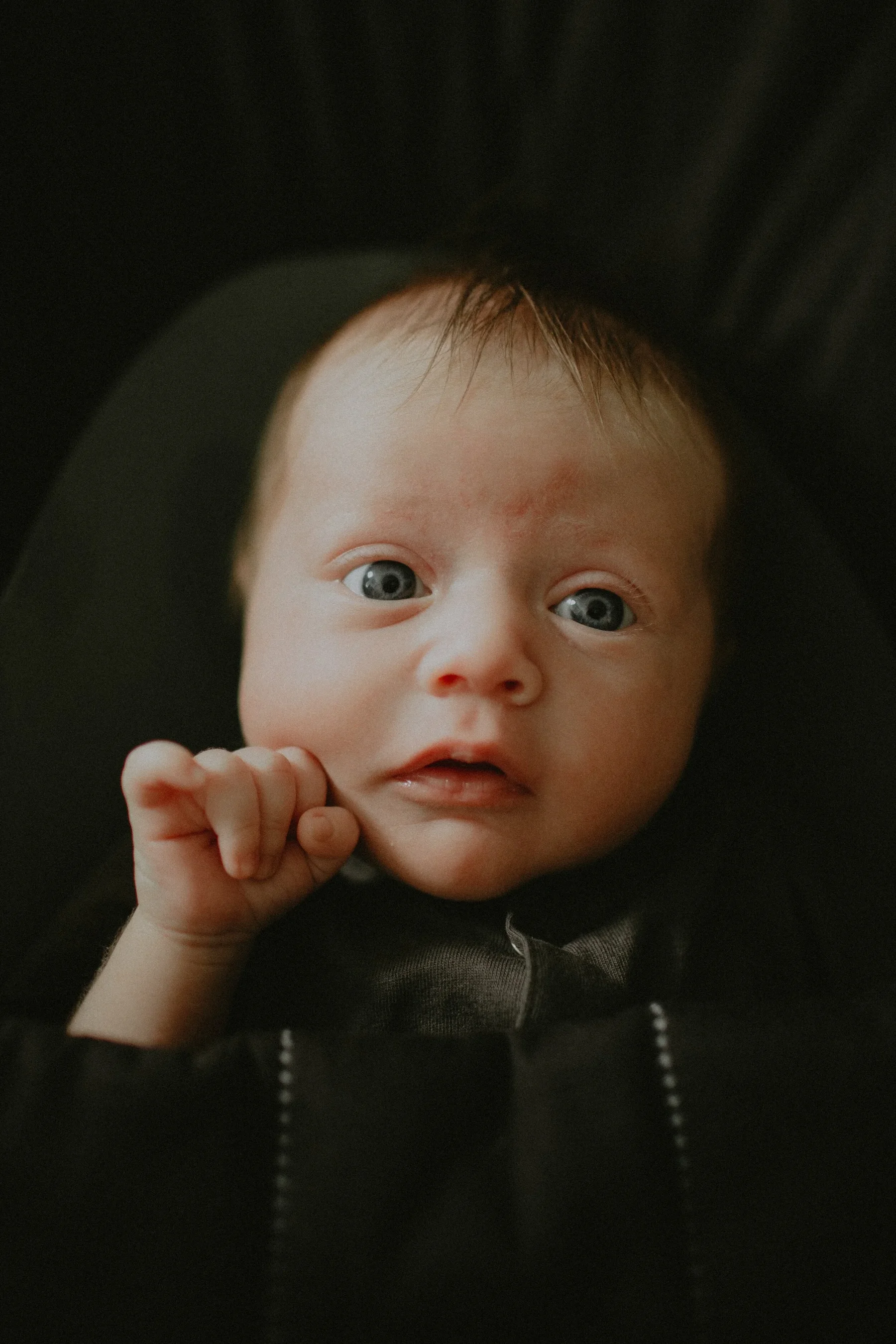 Close-up of a baby with blue eyes and light brown hair, wearing a dark shirt, resting on a black surface.