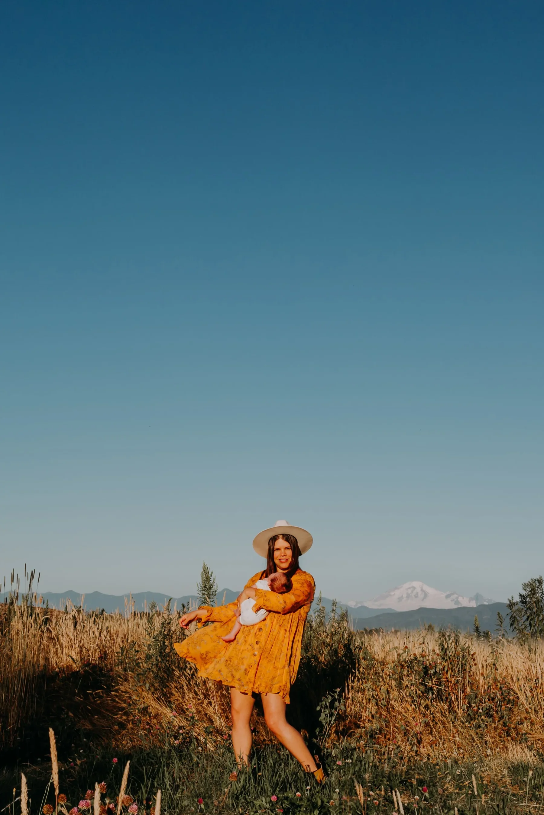 Woman in yellow dress and white hat holding a baby in a field with mountains and snow in the background.